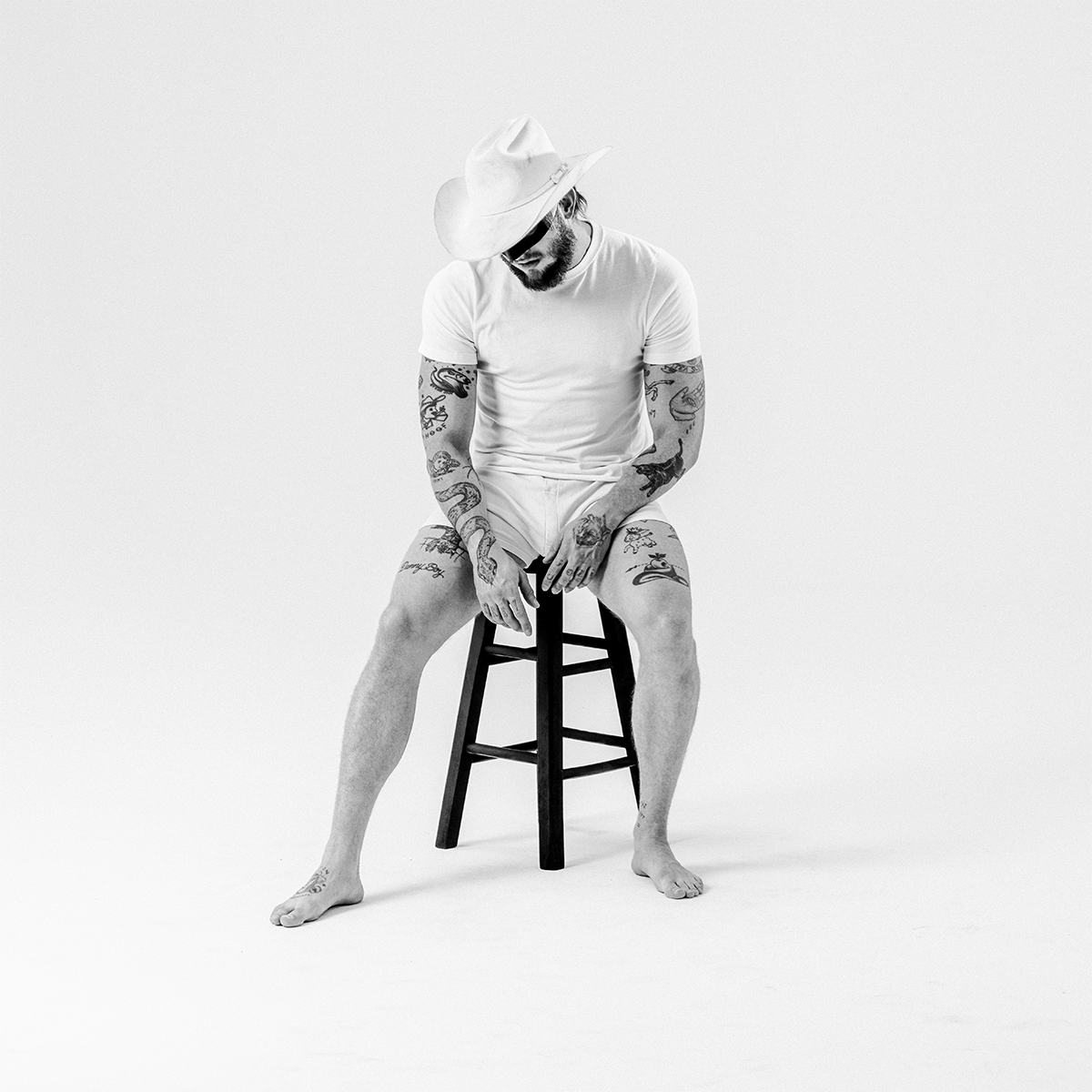 A black and white studio photograph of a Orville Peck wearing a white cowboy hat, white t-shirt, and white shorts, sitting on a black stool against a seamless white background.